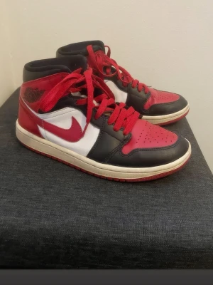 Nike Air Jordan 1 Mid röd/svart/vit - Snygga Nike Air Jordan 1 Mid sneakers i klassisk röd, svart och vit färgkombination. Skorna har läderöverdel, röda skosnören och ikonisk Swoosh-logga på sidan. Perfekta för dig som gillar streetwear och vill sticka ut med en tidlös modell.