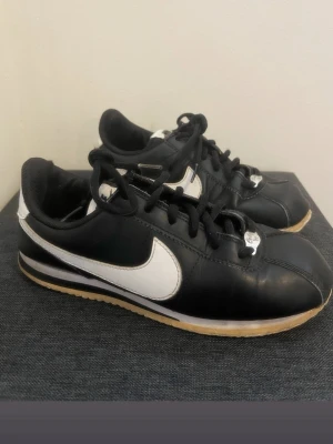 Nike Cortez svartvita sneakers - Klassiska Nike Cortez sneakers i svart och vitt med läderöverdel, svart snörning och vit sula med beige yttersula. Den ikoniska vita swoosh-loggan på sidan och retroinspirerad design gör dem till ett måste för sneakerheads.