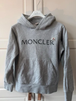 Grå Moncler hoodie med logga - Snygg grå hoodie från Moncler med stor svart logga på bröstet och klassisk Moncler-patch på vänster sida. Hoodien har huva och en stor magficka framtill. Tillverkad i mjukt material som känns skönt mot huden. Perfekt för en avslappnad och trendig look.