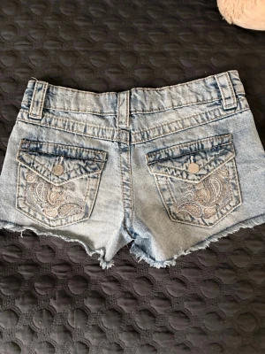 Gina tricot jeansshorts med broderi stl 146 - Snygga ljusblå jeansshorts med slitna kanter och broderade detaljer på bakfickorna. Klassisk femficksmodell med knapp och dragkedja framtill. Perfekta för sommardagar och festivalhäng.