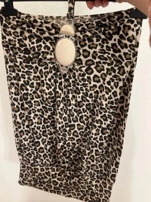 Kort leopardmönstrad blus med smycken - En snygg, beskuren blus i leopardmönster med halterneck och juvelbroderi framtill. Dess eleganta beige och svarta design är perfekt för dig som söker en distinkt look. Tillverkad av ett mjukt, stretchigt tyg för maximal komfort.
