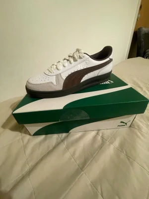 Puma vita och bruna sneakers - Snygga vita sneakers från Puma med bruna detaljer och svart sula. Klassisk låg modell med perforerad tå och ikonisk Puma-logga på sidan. Ovandelen är i läder och mocka, vilket ger en stilren och sportig look. Perfekta för dig som gillar streetwear och enkel stil.