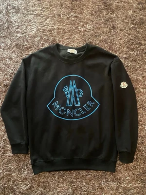 Svart Moncler sweatshirt med blå logga - Snygg svart sweatshirt från Moncler med stor blå logga framtill och klassisk rund hals. Tröjan har ribbade muddar vid ärmslut och nederkant samt Moncler-märke på ena ärmen. Perfekt för en trendig och avslappnad stil.