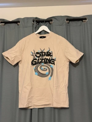 Beige Star Gazing t-shirt  - Cool beige t-shirt med grafiskt tryck “Star Gazing” i svart och blått på framsidan. Kortärmad modell i mjuk bomull med spiral och stjärnor som detaljer. Liten logotyp på ryggen upptill. Perfekt för dig som gillar unika prints och streetstyle.