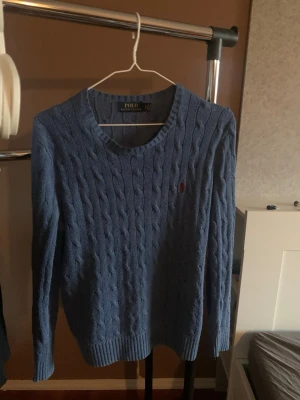 Blå kabelstickad tröja från Polo Ralph Lauren - Kabelstickad blå tröja från Polo Ralph Lauren med rund halsringning och röd broderad logga på bröstet. Tröjan har långa ärmar och ribbstickade muddar vid ärmslut och nederkant. Perfekt för en stilren och klassisk look.