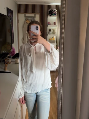 Vit plisserad blus med knytband - Säljer en vit, luftig blus med plisserat tyg och knytband vid halsen. Blusen har långa, vida ärmar och en avslappnad passform. Perfekt till jeans eller kjol för en trendig look.