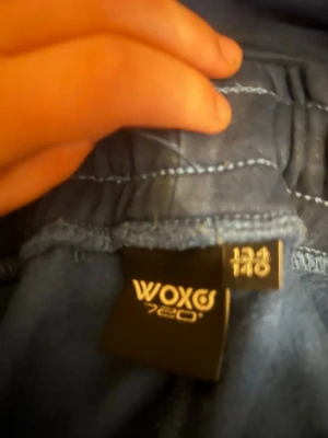 Blå mjukisbyxor från Woxo 720 - Mörkblå mjukisbyxor från Woxo 720 med tryckt vit logga på ena benet. Byxorna har resår i midjan med dragsko och muddar vid bensluten. Materialet ser ut att vara mjukt och bekvämt, perfekt för chill dagar eller träning. Snygg och enkel design som funkar till det mesta.