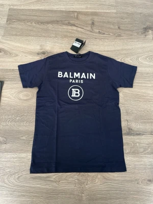 Marinblå Balmain Paris t-shirt - Snygg marinblå t-shirt från Balmain Paris med vit logga och tryck framtill. Klassisk rund halsringning och korta ärmar. Perfekt för en trendig och stilren look.