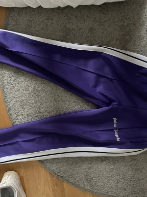 Lila mjukisbyxor från Palm Angels - Snygga lila mjukisbyxor från Palm Angels med vita sidoränder och logga på låret. Byxorna har en sportig look med raka ben och elastisk midja. Perfekta för en avslappnad streetwear-stil.