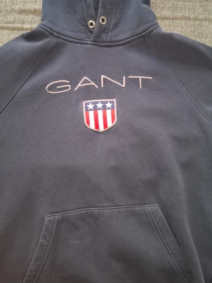 Mörkblå hoodie från GANT med logga - Säljer en mörkblå hoodie från GANT med broderad logga och amerikanskinspirerat märke på bröstet. Klassisk känguruficka framtill och huva med dragsko. Perfekt för dig som gillar stilrena och sportiga plagg.