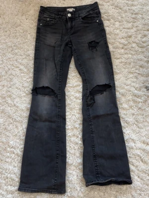 Svarta bootcut jeans med slitningar - Säljer ett par svarta bootcut jeans med egengjorda stora slitningar över knäna och en mindre vid fickan. Jeansen har knappgylf och utsvängda ben. Perfekta för en edgy och avslappnad stil.