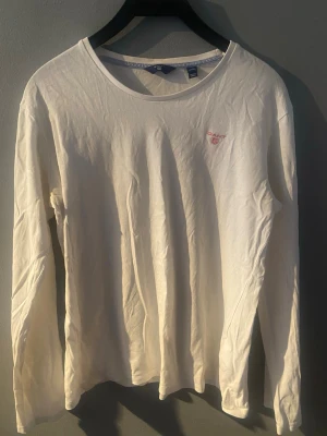 Vit långärmad topp från Gant, strl 170 - Klassisk vit långärmad topp från Gant med rund halsringning och diskret broderad logga i rosa på bröstet. Tillverkad i mjuk bomul. Perfekt basplagg.