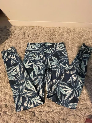 Mönstrade leggings med blått bladmotiv - Snygga leggings med tropiskt bladmotiv i blått, vitt och turkost. Hög midja och stretchigt material som sitter skönt. Perfekta för dig som gillar att sticka ut med coola mönster. Passar till både träning och chill.