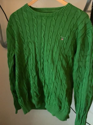 Grön kabelstickad tröja från Gant - Snygg grön kabelstickad tröja från Gant med rund halsringning och diskret broderad logga på bröstet. Tröjan har långa ärmar och ribbade muddar vid ärmslut och nederkant. Perfekt för en stilren och avslappnad look.