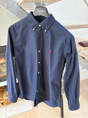 Mörkblå skjorta från Polo Ralph Lauren - Klassisk mörkblå skjorta från Polo Ralph Lauren med button-down krage och vit knappstängning. Skjortan har lång ärm och röd broderad logga på bröstet. Perfekt för en stilren och avslappnad look.