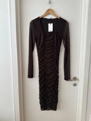 Brun långärmad klänning från BikBok - Snygg brun långklänning från BikBok med långa ärmar i mesh och rynkade detaljer längs hela klänningen. Fyrkantig ringning och figurnära passform ger en trendig look. Perfekt för dig som vill sticka ut med stil.