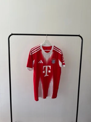 Bayern München matchtröja Adidas röd - Röd och vit Bayern München matchtröja från Adidas med klubbmärke och sponsortryck på bröstet. Tröjan har klassiska Adidas-ränder på axlarna och rund hals. Tillverkad i lätt och ventilerande material, perfekt för fotbollsträning eller match.