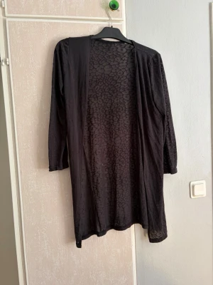 Svart lång cardigan med mönster - Svart lång cardiganjacka med diskret djurmönster. Öppen framtill utan knäppning, långärmad och i tunt, mjukt material. Perfekt att slänga över en t-shirt eller topp för en chill look.