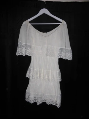 Vit omlottklänning med spetsdetaljer - Superfin vit kortklänning i boho-stil med volanger och broderade spetsdetaljer. Klänningen har offshoulder-modell och v-ringning, samt vackra blommiga broderier längs kanterna. Perfekt för sommarens alla tillfällen.
