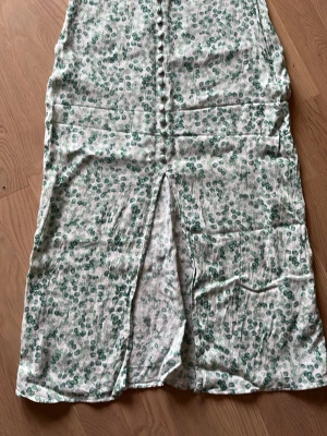Grönblommig långkjol med knappar - Långkjol med grönt blommönster på vit botten. Kjolen har dekorativa knappar framtill och en slits som ger en snygg detalj. Dragkedja vid sidan, köpte den själv begagnad men lite för liten för mig.