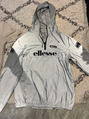 Reflekterande vindjacka från Ellesse - Snygg reflekterande vindjacka från Ellesse med stor logga på bröstet och ärmarna. Jackan har huva med dragsko, halv dragkedja framtill och elastiska muddar. Perfekt för dig som vill sticka ut med en sportig och modern look. Den har synliga tecken på användning så jag säljer jackan på ett mer lämpligt pris! 