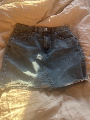 Blå jeanskjol med rå kant - Säljer en blå jeanskjol i klassisk modell med fem fickor och rå, fransig kant nedtill. Kjolen har normal passform och är gjord i denim med en snygg slits framtill. Perfekt till sommaren och enkel att styla.