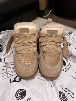Beige UGG sneakers med fuskpäls - Snygga beige sneakers från UGG med tjocka snören och fuskpäls på insidan. Skorna har en mix av mocka och mesh-material, samt en robust sula. Perfekt för dig som vill ha både stil och komfort.