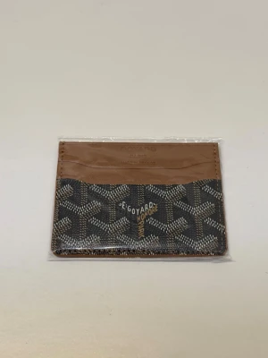Korthållare från Goyardt Saint Sulpice  - Säljer en tidlös accessoar som är både lyxig och praktisk. Skick: Helt ny(ligger fortfarande i sin skyddsplast). Material: Äkta läder. Färg: Svart mönster med bruna (tan) läderdetaljer.Design: 4 kortfack (2 på varje sida) samt ett större fack i mitten för sedlar eller kvitto. Extremt smidig profil som passar perfekt i fickan.Perfekt som present till dig själv eller någon annan. Kan skickas spårbart eller i Stockholm. Nypris ligger högt och modellen är ofta slutsåld i butik.