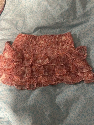 Rosa volangkjol med paisleymönster - Supersöt kort kjol från Gina i rosa med paisleyinspirerat mönster och volanger. Kjolen har bred smock i midjan för en bekväm passform och är gjord i ett lätt och luftigt material, perfekt för varma dagar.