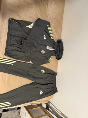 Real Madrid träningsset från Adidas - Säljer ett grått träningsset från Adidas med Real Madrid-logga. Setet består av en jacka med huva och matchande byxor, båda med ljusgula detaljer och klassiska Adidas-ränder. Perfekt för dig som vill representera Real Madrid på träningen.