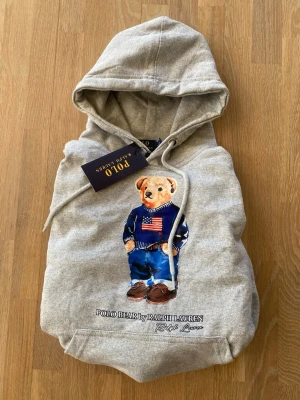 Grå hoodie Polo Bear Ralph Lauren - Säljer en grå hoodie från Polo Ralph Lauren med den ikoniska Polo Bear-trycket framtill. Hoodien har dragsko i huvan och en klassisk känguruficka. Mjuk bomullsblandning som är perfekt för en chill och stilren look.