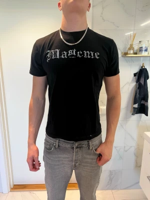 Svart t-shirt från MASTEME - Svart t-shirt från MASTEME med tryck i vitt framtill och en stor grafisk illustration på ryggen. T-shirten har korta ärmar och en normal passform. Materialet är mjuk bomull, perfekt för en avslappnad streetwear-look. Vissa pärlor har lossnat