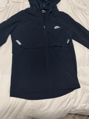 Svart Nike hoodie med dragkedja - Snygg svart hoodie från Nike med hel dragkedja framtill och vit logga på bröstet. Utrustad med huva och reflekterande detaljer vid fickorna. Perfekt för en sportig och avslappnad stil.