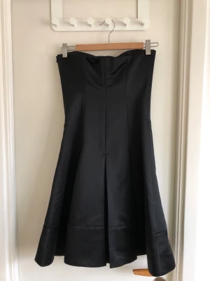 Svart axelbandslös klänning MANGO SUIT - Elegant svart axelbandslös långklänning med utsvängd kjol och markerad midja. Klänningen har en stilren och tidlös design i ett glansigt material som ger en lyxig känsla. Perfekt för dig som vill sticka ut med en klassisk look.