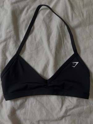 Svart sporttop från Gymshark - En svart sporttop från Gymshark med smala axelband och V-ringning. Mjuk och stretchig modell utan byglar, perfekt för en sportig look. Diskret logga framtill i vitt. Materialet känns följsamt och bekvämt mot huden.