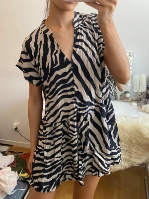 Zebramönstrad kort klänning H&M - Snygg kort klänning från H&M med zebramönster i svart och vitt. Klänningen har v-ringning, korta ärmar och en lös passform med lätt volang nedtill. Perfekt för dig som gillar trendiga och djärva mönster.
