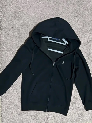 Svart hoodie från Ralph Lauren - Svart hoodie med dragkedja från Ralph Lauren. Klassisk design med huva, fickor framtill och den ikoniska vita broderade loggan på bröstet. Perfekt för en chill och stilren look. Mjuk bomullsblandning och ribbade muddar.