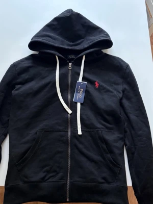 Svart hoodie från Polo Ralph Lauren - Svart hoodie med dragkedja från Polo Ralph Lauren. Klassisk design med vit dragsko, huva och två fickor framtill. Röd broderad logga på bröstet. Mjuk bomullsblandning som är skön att ha på sig.
