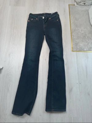 Mörkblå bootcut jeans från True Religion - Snygga mörkblå bootcut jeans från True Religion med klassiska kontrastsömmar och detaljerade bakfickor med lock och knappar. Jeansen har låg midja och silverfärgade nitar. Perfekta för en trendig och avslappnad look.