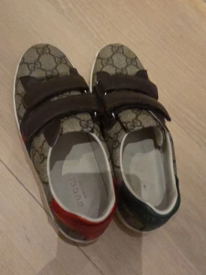 ÄKTA Gucci sneakers med monogram och kardborre - Snygga Gucci sneakers med klassiskt beige GG-monogram, bruna kardborreband och vita sulor. Skorna har gröna och röda detaljer på sidorna samt olika färg på hälen – en röd och en grön. Perfekta för dig som vill sticka ut med lyxig streetstyle.