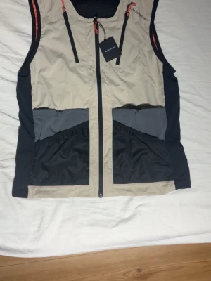 Beige och svart funktionsväst från Klättermusen - Snygg funktionsväst från Klättermusen i beige och svart med grå detaljer. Västen har dragkedja framtill, stora fickor och justerbara snoddar baktill. Perfekt för dig som gillar outdoor och vill ha en praktisk men stilren look.