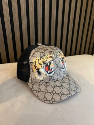 GG keps med tigertryck - Snygg keps från Gucci med klassiskt GG-mönster och två stora tigerhuvuden framtill. Framsidan är beige med brunt mönster, baksidan är svart mesh och justerbar rem med grönt och rött band. Perfekt för dig som vill sticka ut med en lyxig accessoar.