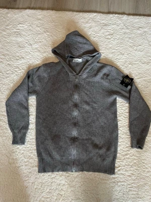Grå hoodie från Stone Island - Säljer en grå ribbstickad hoodie från Stone Island med dragkedja framtill och ikonisk svart-gul patch på ärmen. Hoodien har långa ärmar och en skön huva. Perfekt för dig som gillar streetwear och vill ha något stilrent men ändå med edge.