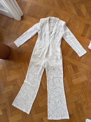 Vit spetsjumpsuit med kavajkrage - Supersnygg vit jumpsuit i spets med långa ärmar och kavajkrage. Jumpsuiten har vida ben, fickor framtill och stängs med en knapp. Perfekt för dig som vill sticka ut med en unik och trendig look.