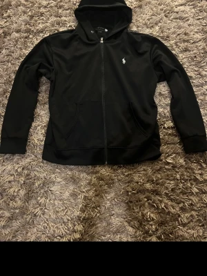 Svart hoodie från Ralph Lauren - Svart hoodie med dragkedja från Ralph Lauren. Klassisk design med huva, två fickor framtill och den ikoniska lilla vita loggan på bröstet. Perfekt för en avslappnad och stilren look.