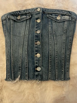 Denim korsett från Zara med knappar - Cool denim korsett från Zara i blått med silverfärgade knappar framtill och två dekorativa fickor. Korsetten har rå fransig nederkant och markerade sömmar som ger en edgy look. Perfekt för att styla med jeans eller kjol.