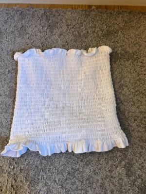 Vit bandeau tubtopp med volang - Söt vit bandeau tubtopp med smockad struktur och volangkant upptill och nedtill. Perfekt för varma dagar och enkel att matcha med jeans eller kjol. Toppens material känns mjukt och stretchigt.