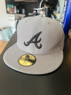 Grå New Era 59Fifty keps Atlanta - Snygg grå keps från New Era 59Fifty med Atlanta Braves-logga broderad framtill i svart. Klassisk flat peak och broderade ventilationshål. Kepsen är i fast storlek och har MLB-logga baktill. Perfekt för dig som gillar streetwear.