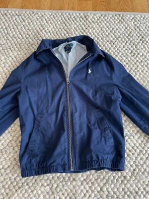 Marinblå vindjacka från Polo Ralph Lauren - Snygg marinblå vindjacka från Polo Ralph Lauren med dragkedja framtill, två sidofickor och klassisk vit logga på bröstet. Jackan har elastisk nederkant och ärmavslut samt randigt innerfoder. Perfekt för en stilren och sportig look.