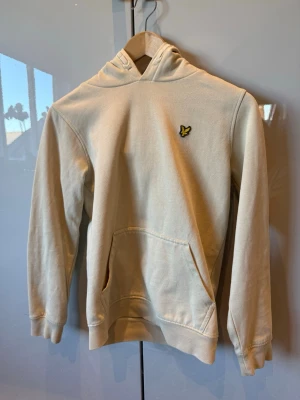 Beige hoodie från Lyle & Scott - Snygg beige hoodie från Lyle & Scott med klassisk gul logga på bröstet. Hoodien har huva, känguruficka och ribbade muddar vid ärmslut och nederkant. Tillverkad i mjukt bomullsmaterial som är skönt att bära.